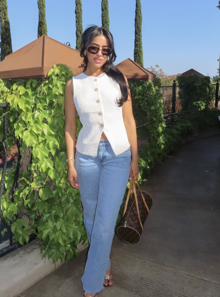 Woman wearing a structured white vest with high-waisted jeans, carrying a statement bag, and wearing sunglasses.